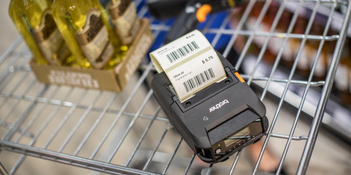 A Brother portable label printer with a barcode label next to olive oil stock