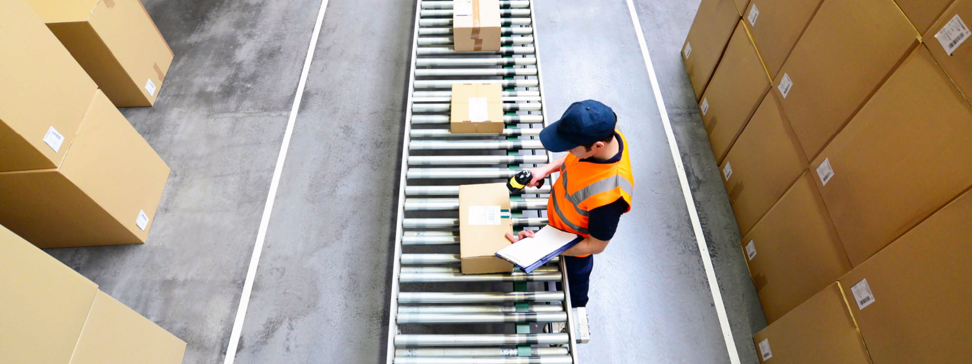 A warehouse worker placing labels on packages on a conveyor belt with stock piled up around.