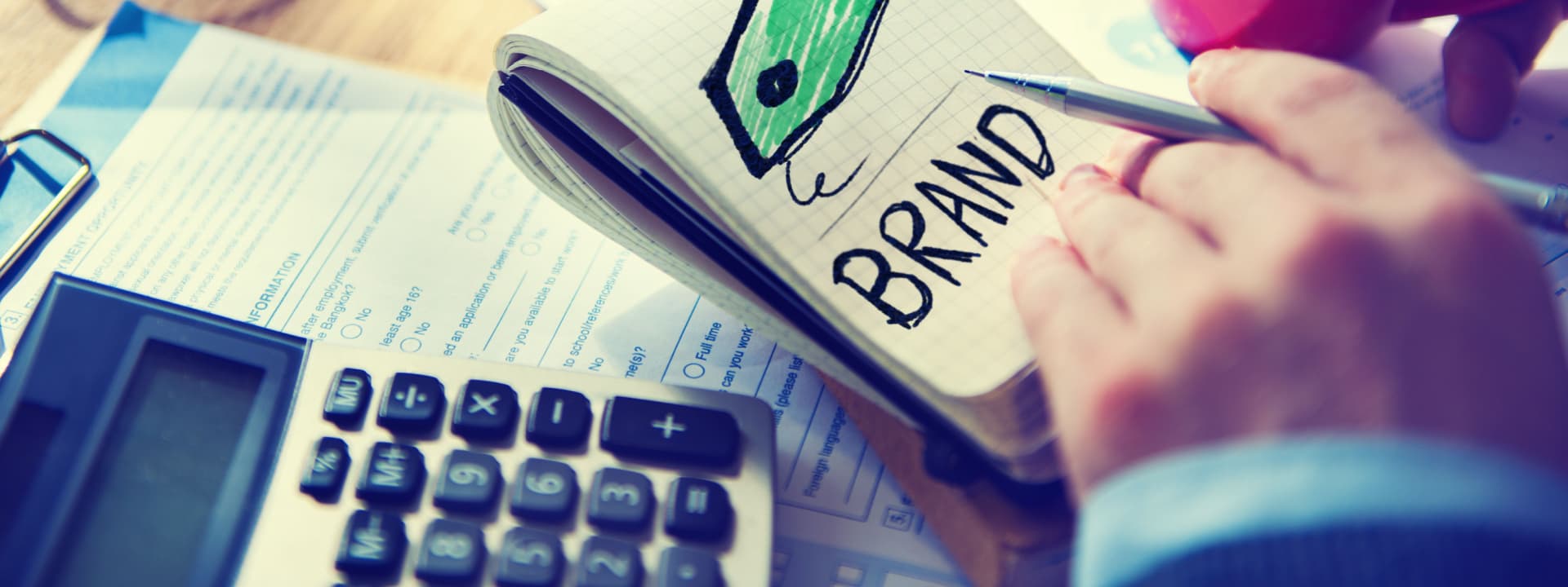 Person's hand with a notepad and brand written on it next to documents and a calculator