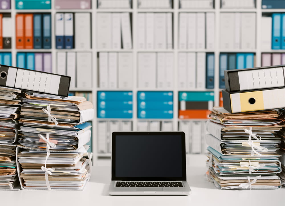 A laptop next to a stack of papers and filing cabinets