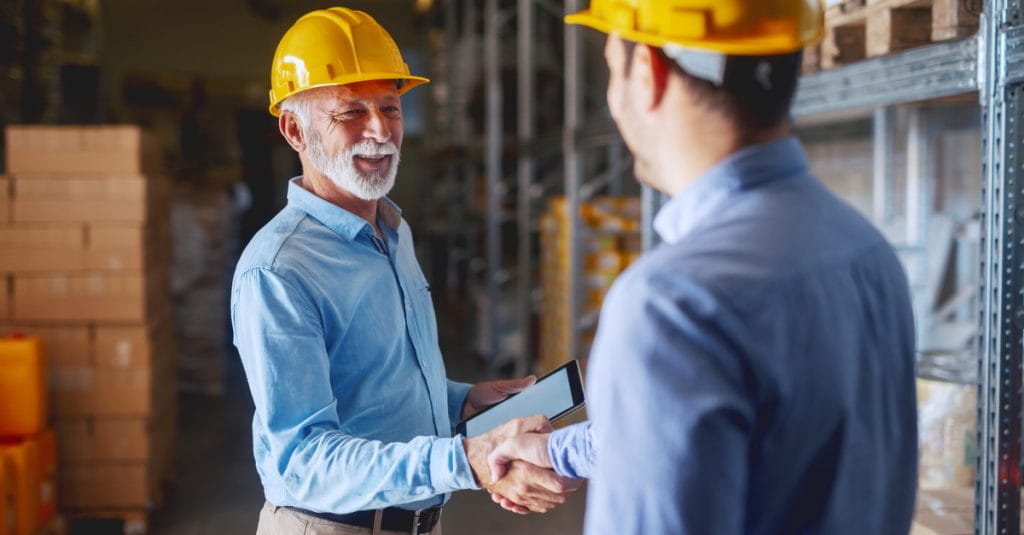 Two warehouse workers with hard hats shaking hands next to inventory in shelves.