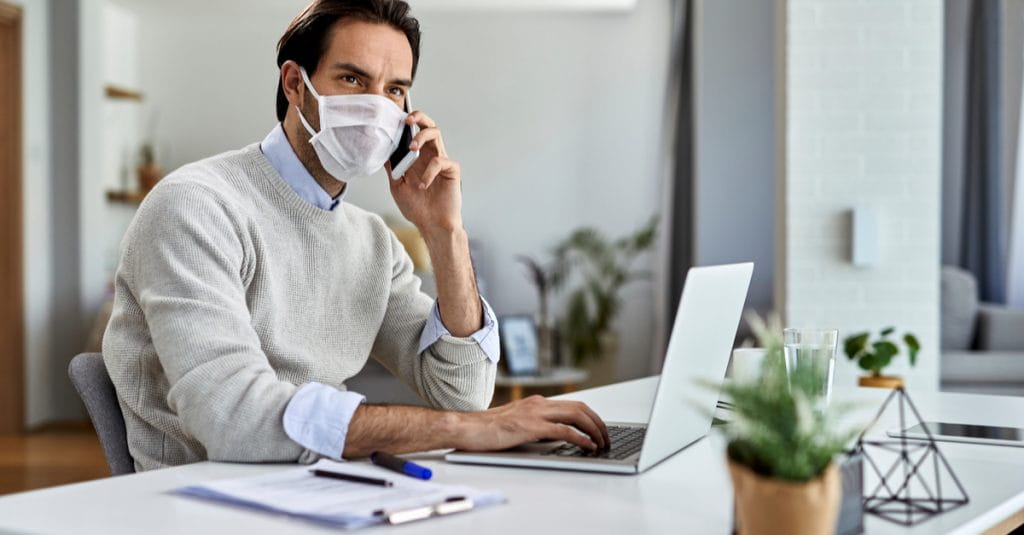 A masked man working from home on his phone in front of a laptop.