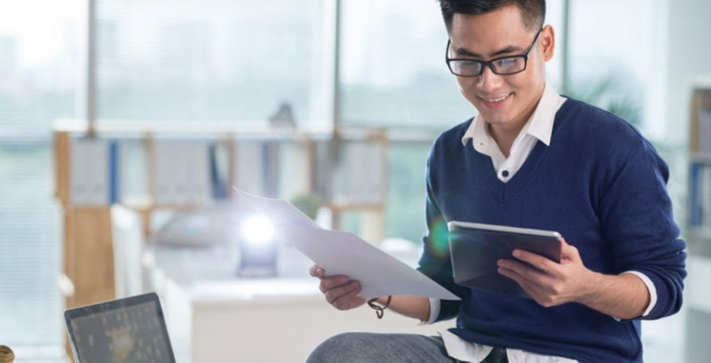 A man working from home holding up a printed document next to his laptop.