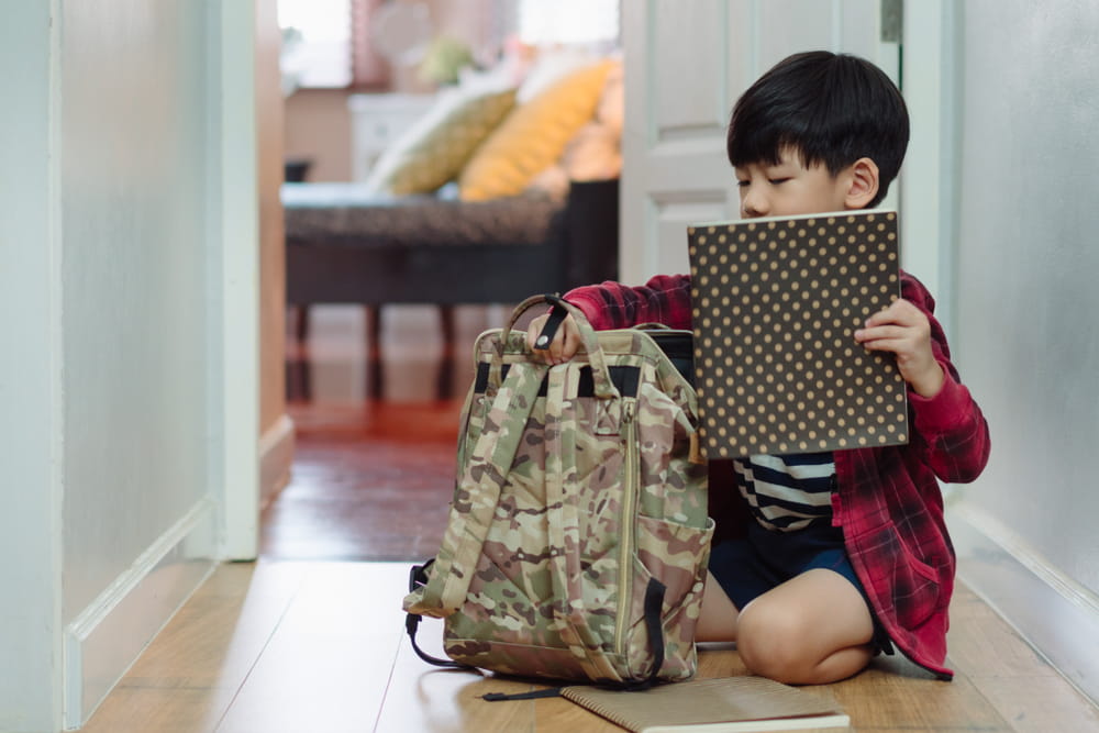 A child sitting down next to his backpack with a notebook in hand.