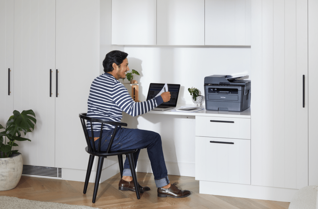 A man sitting at his home office next to a printer and laptop printing documents.