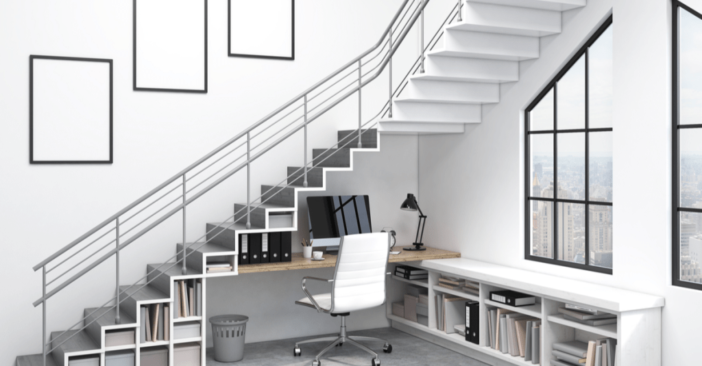 A study nook positioned underneath a white staircase with computer, desk and chair. 