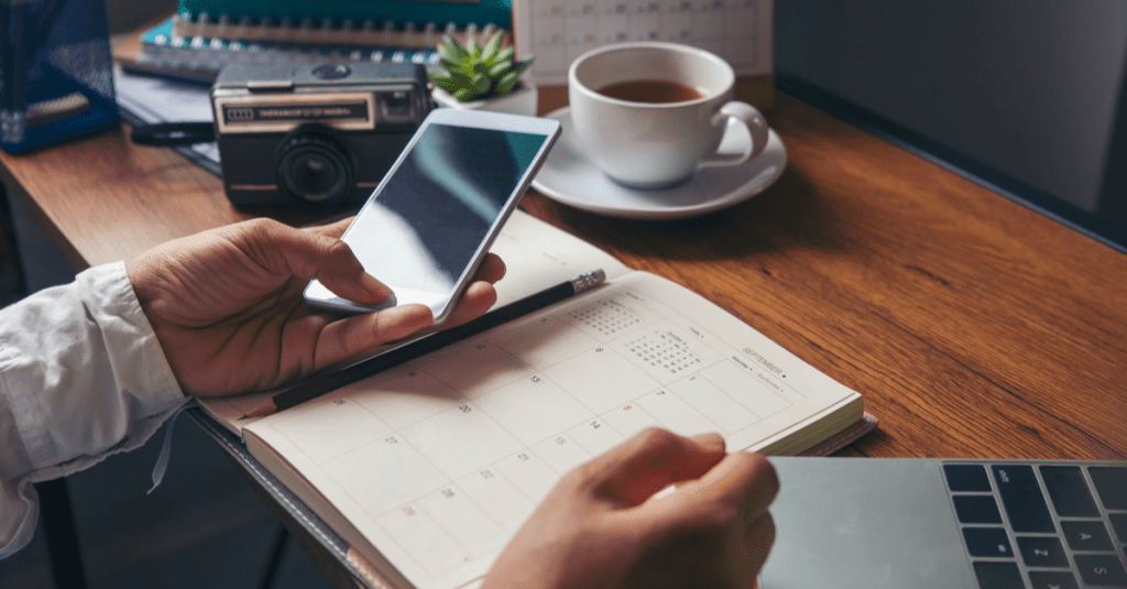 A man on his smartphone at his desk with a calendar and cup of coffee. 