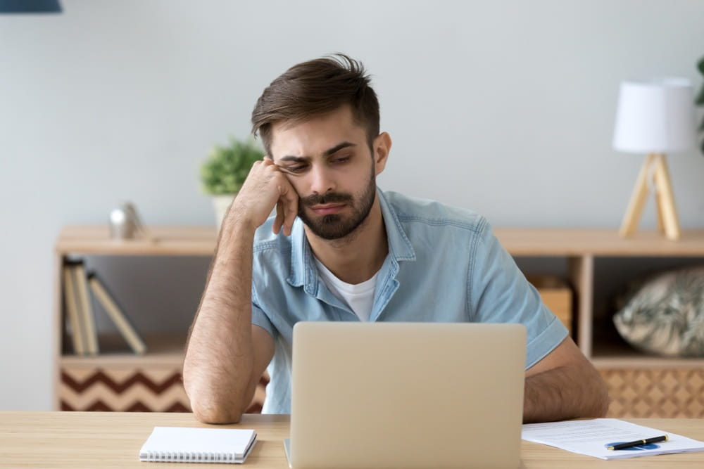 A disengaged male worker looking unenthused in front of his laptop while working from home.