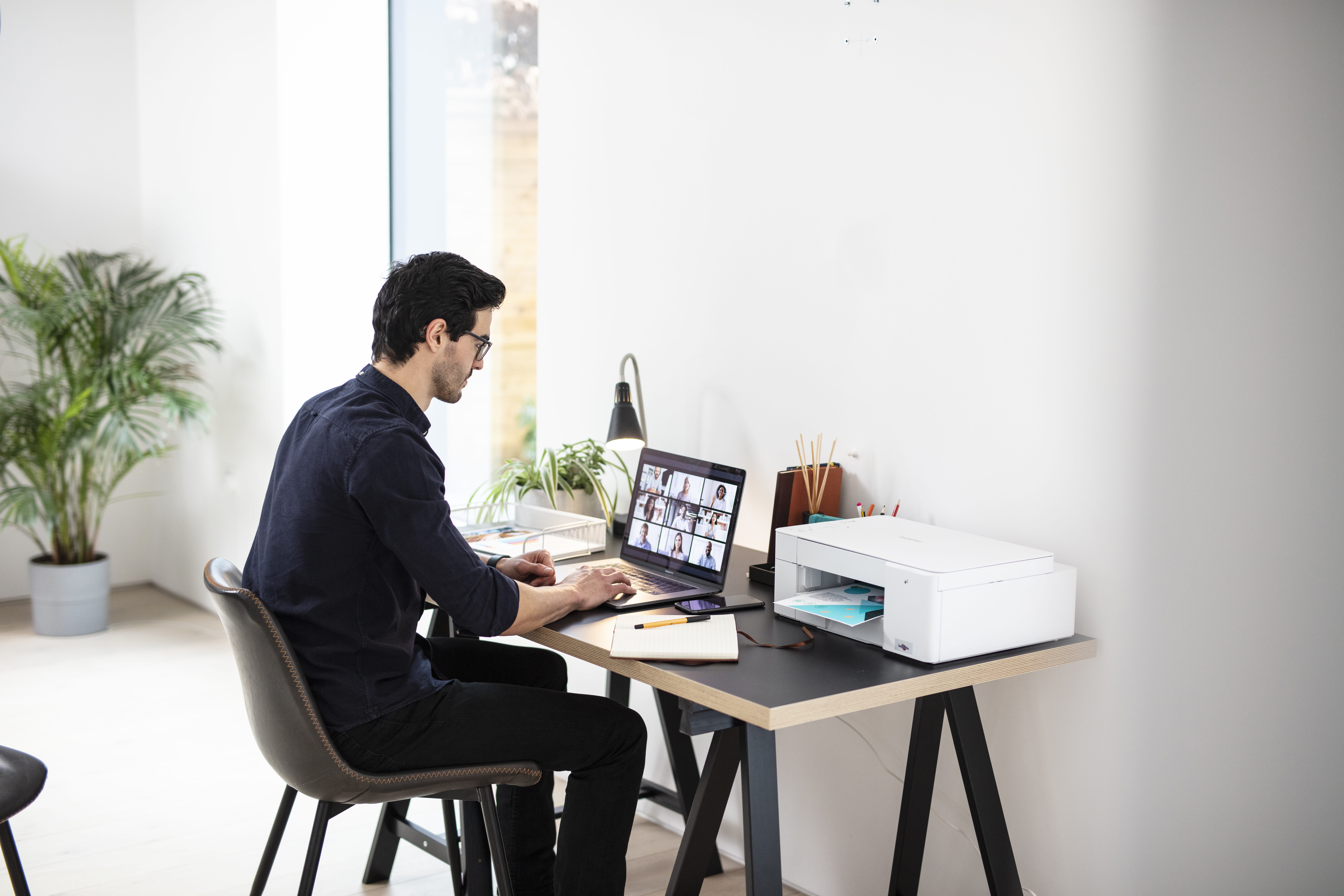 A man sitting at a desk with his laptop open and a DCP-J1200W on the desk