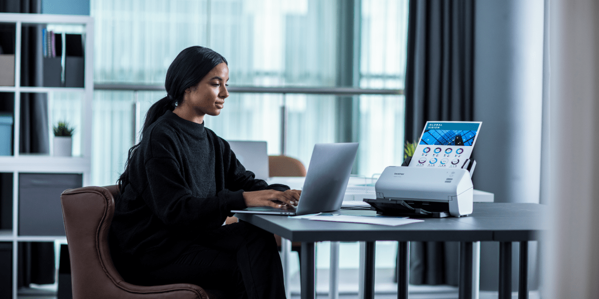 Lady sitting down on a laptop next to a Brother ADS-3100 scanner