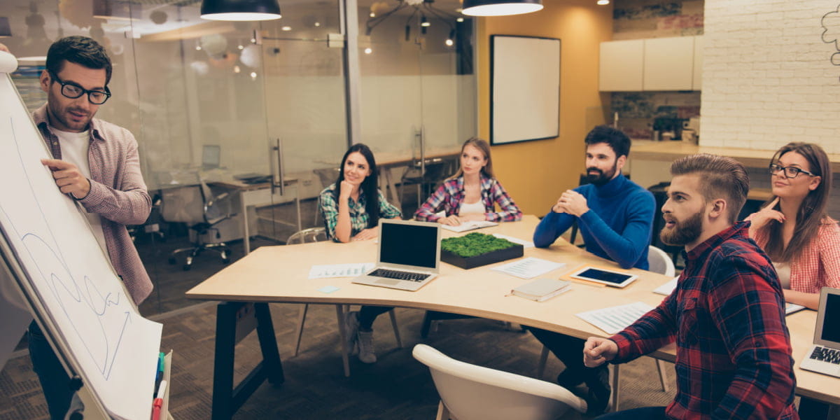 A manager pointing to a whiteboard during a meeting with colleagues