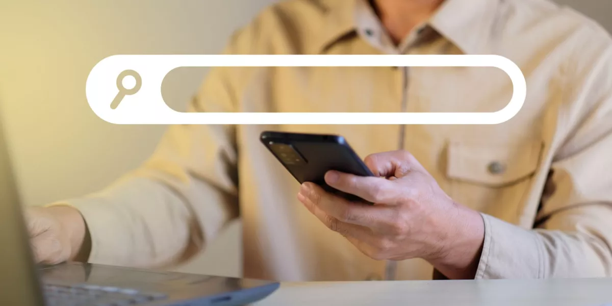 A person on a smartphone using a search engine in front of a laptop