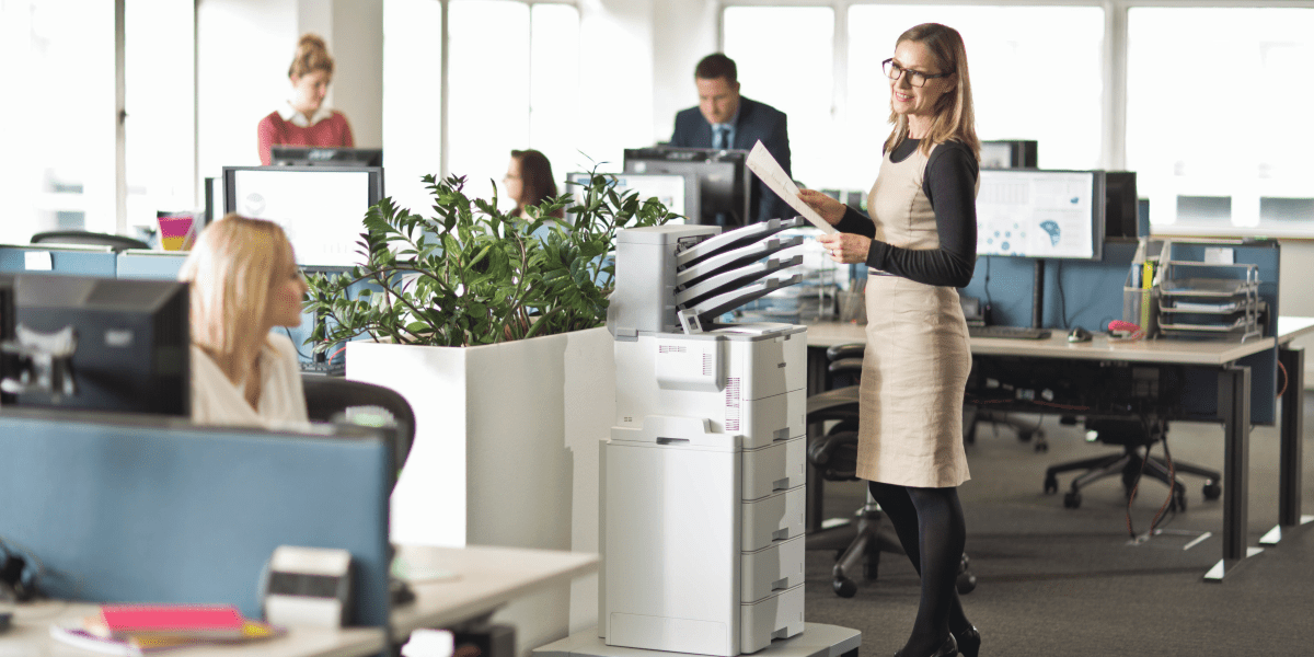 A small office with coworkers around a printing station next to a green plant. 