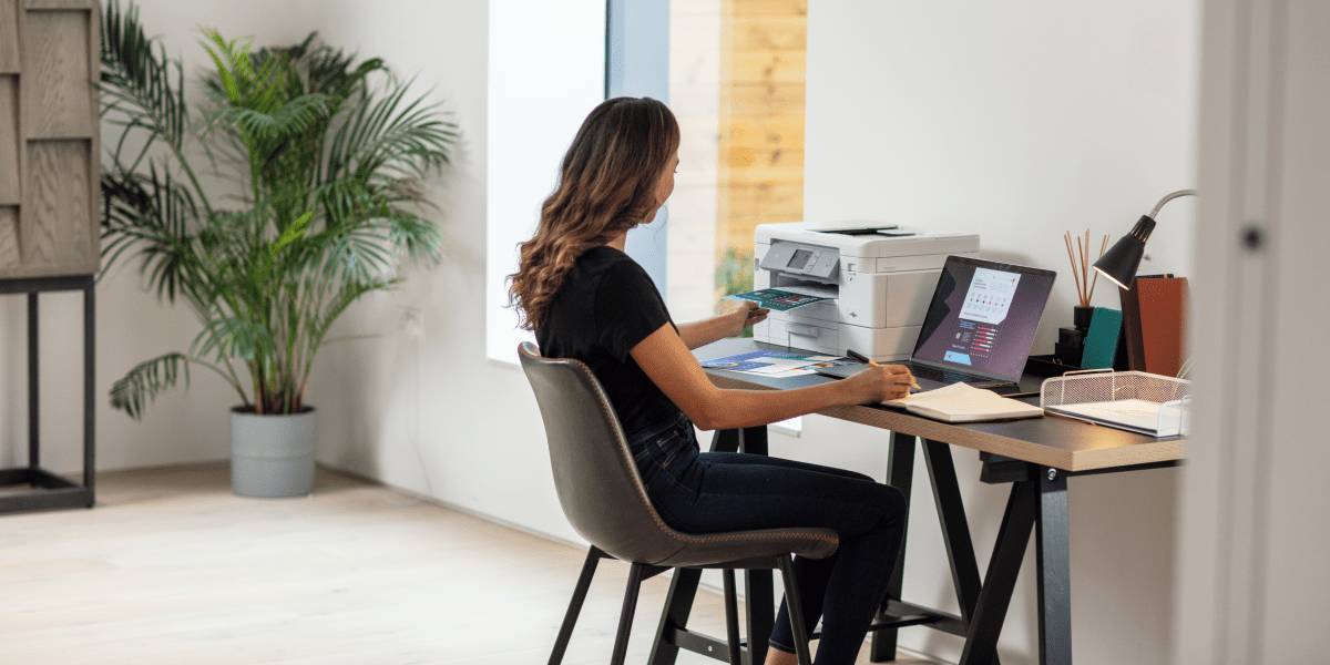 A female business owner working on a laptop from home next to a printer and plant.