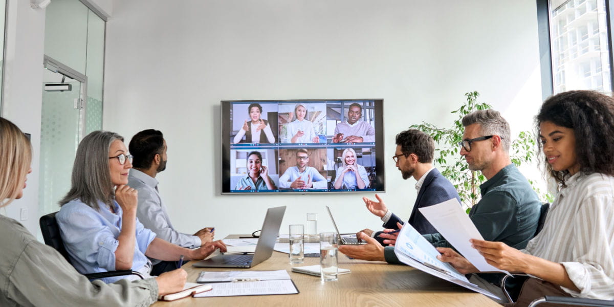 A boardroom of men and women with a screen displaying other colleagues.