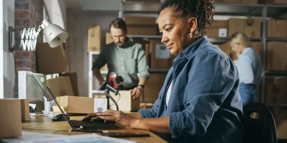 Retail business owner on a laptop in a warehouse with staff looking at inventory on shelves