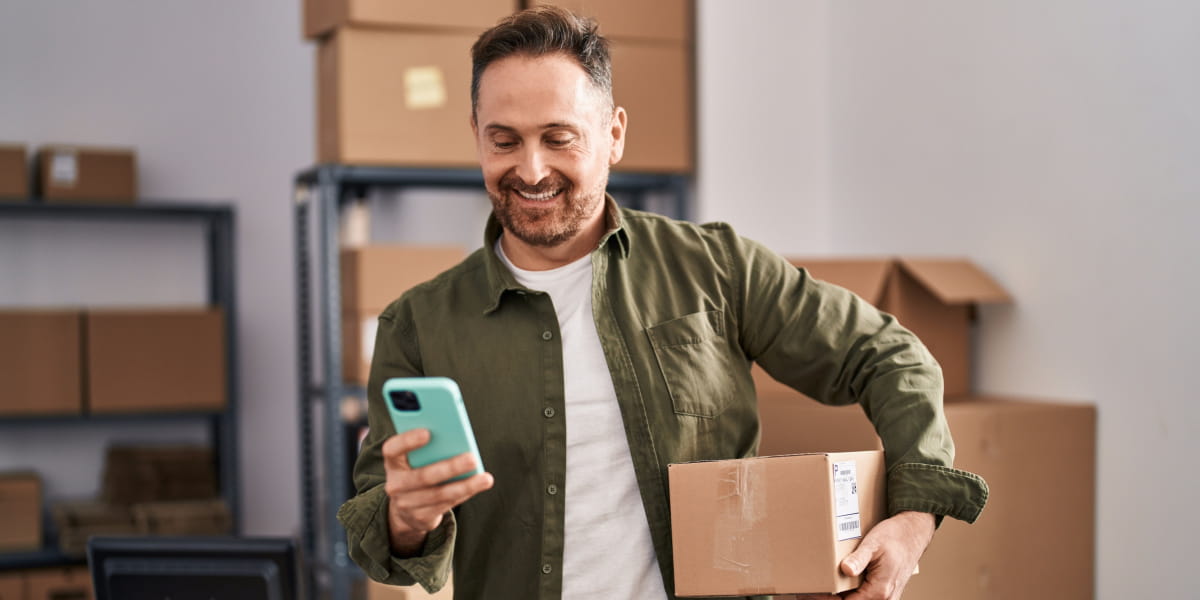A happy male eCommerce business owner smiling at smartphone with a parcel in hand