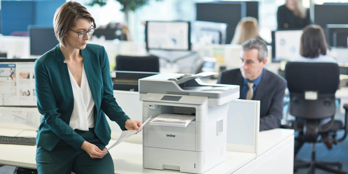 A lady sitting next to a printer in a busy office environment
