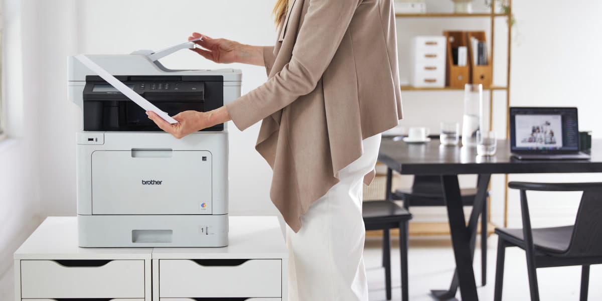 A lady next to a Brother multi-function printer with a sheet of paper in hand
