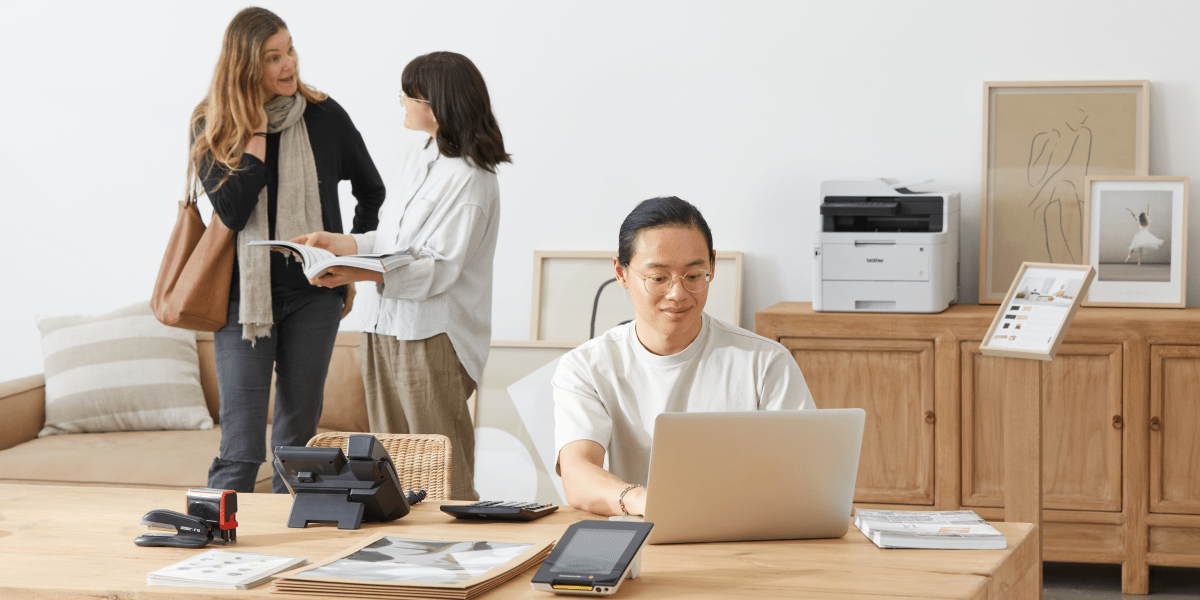 A male sitting down on a laptop with two female colleagues in background next to a printer.