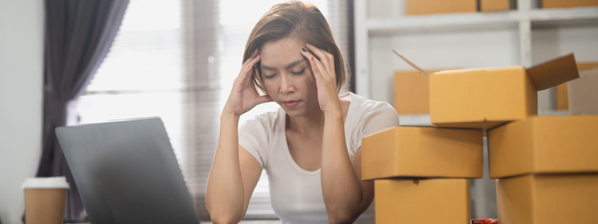A lady ecommerce business owner visibly stressed with packages and a laptop on the desk