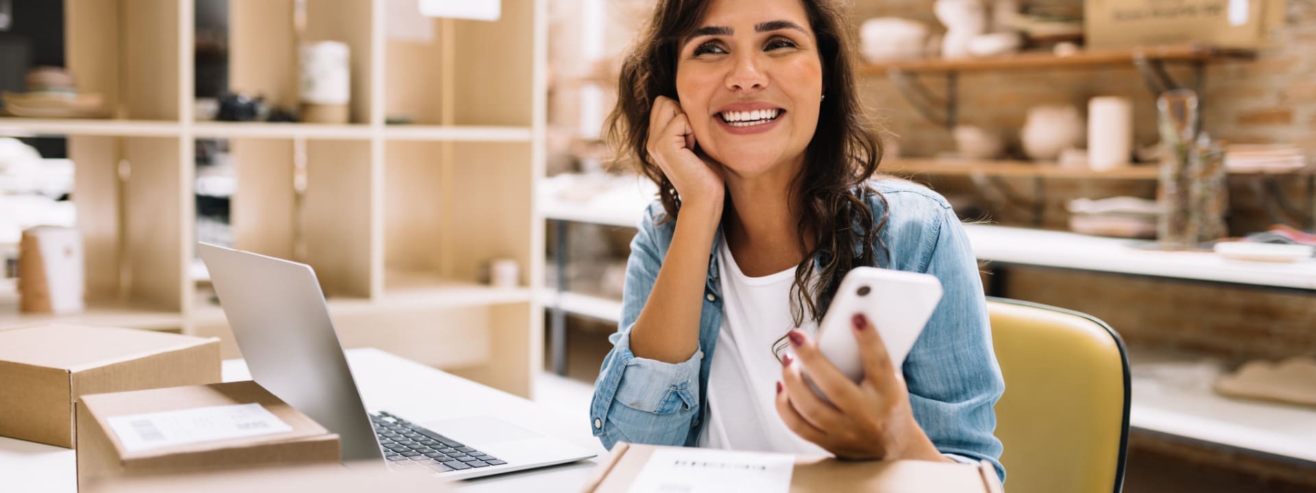 A young ecommerce business owner with a laptop and smartphone next to lots of stock