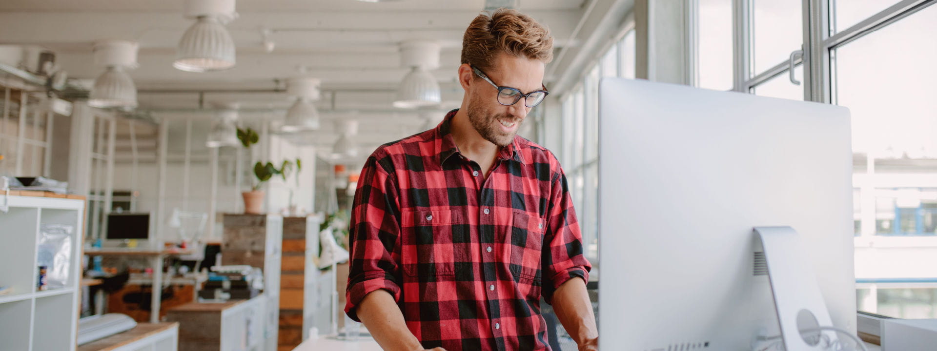 A man standing up in front of his computer with hybrid furniture in the office.
