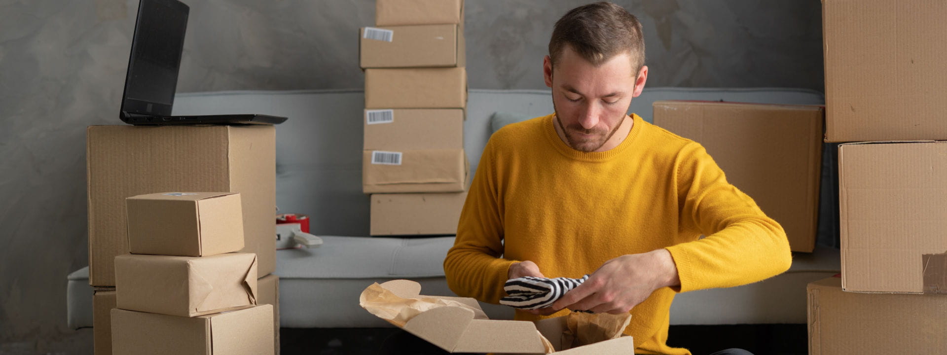 A male ecommerce business owner in a yellow shirt packing boxes for customer orders.