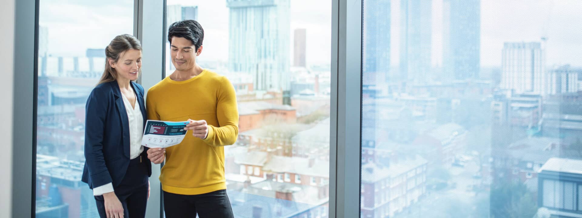 Two co-workers in an office looking at printed documents next to a view of the city