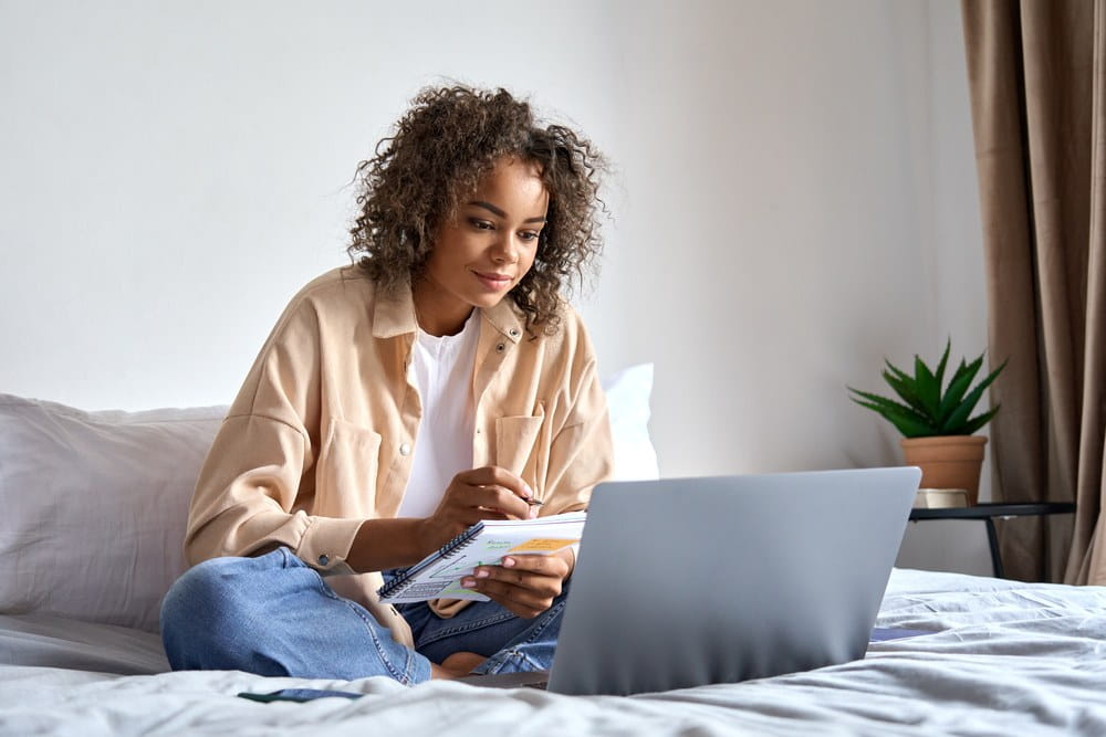 A young lady on her bed working from home with a notepad and laptop.