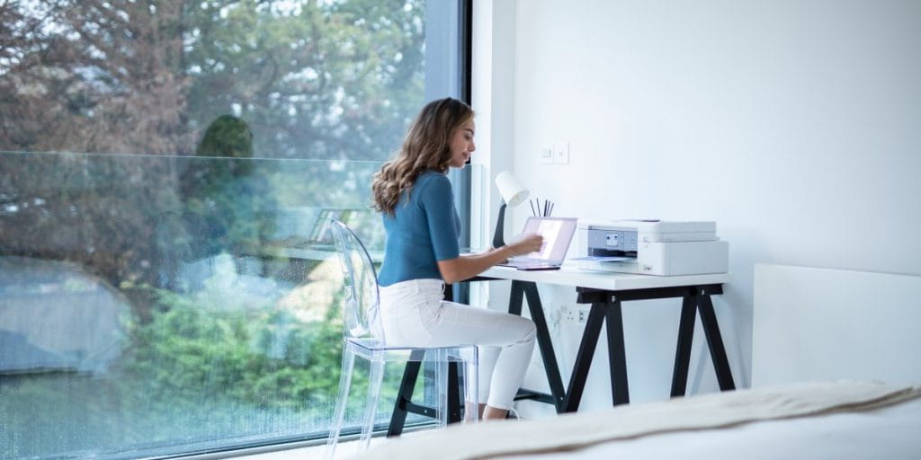 A lady sitting down at a desk in her bedroom with laptop and printer.