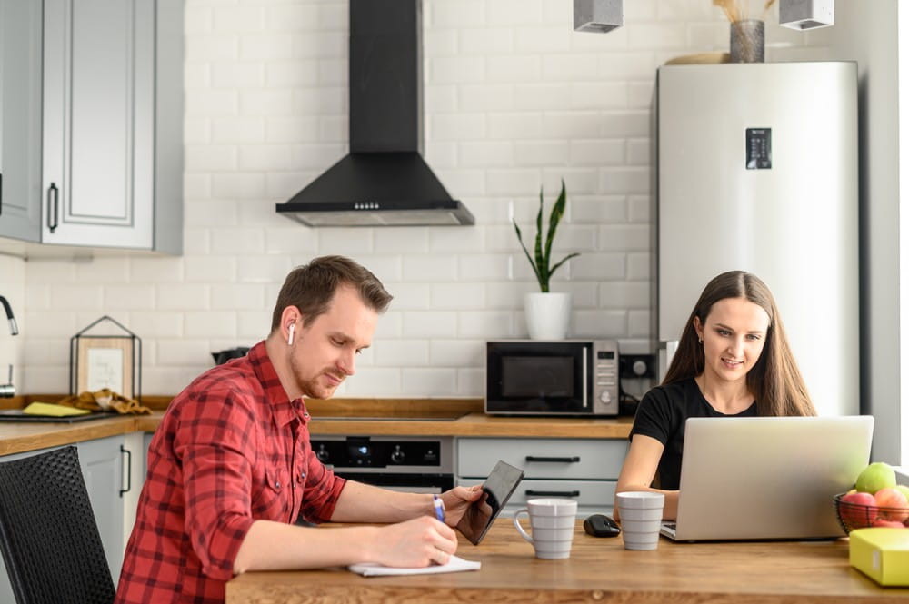 A husband and wife at home working on laptops in the kitchen.