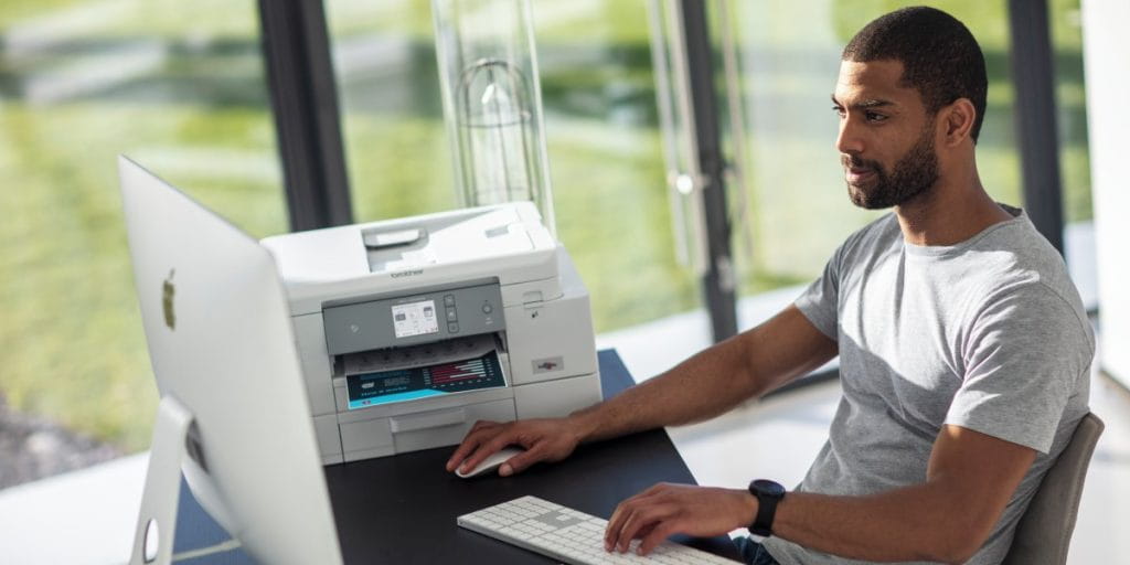 Young male working from home in front of screen and printer on desk with natural light.