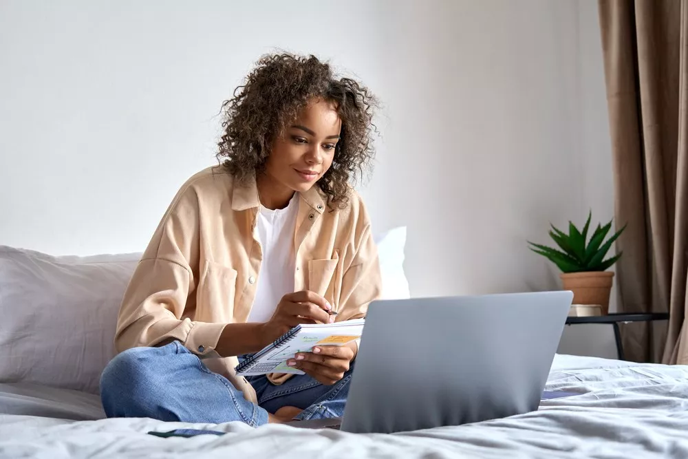 A young lady on her bed working from home with a notepad and laptop.