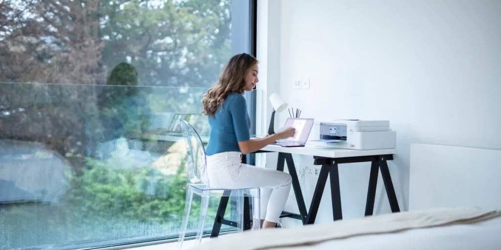 A lady sitting down at a desk in her bedroom with laptop and printer.