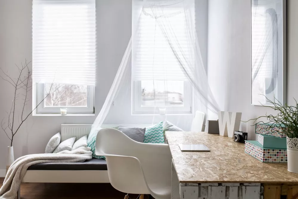 A bedroom home office with open windows and curtains, a desk and chair.