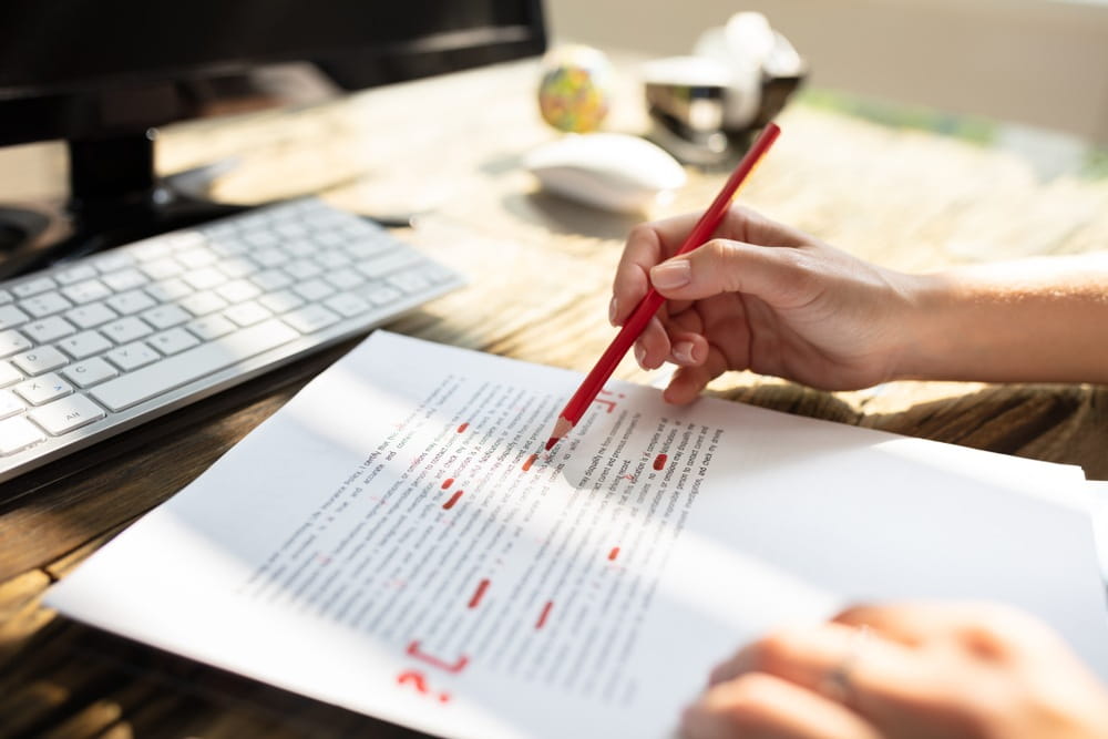 A person editing written copy on paper with a red pencil in front of their laptop.