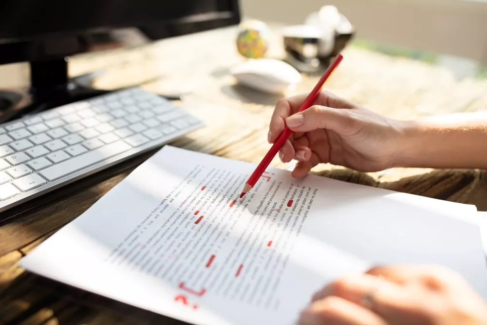 A person editing written copy on paper with a red pencil in front of their laptop.